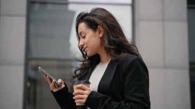 Kız yürüyor, akıllı telefonunu kaydırıyor, gülümsüyor ve içiyor. Koyu saçlı beyaz kadın gri modern binalarda dolaşıyor, akıllı telefonunu kaydırıyor ve kağıt bardaktan sıcak su içiyor..