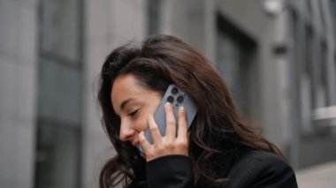 Caucasian businesswoman talking on phone, smiling, looking around, working outdoors on her laptop. Young female working remotely on her computer outdoors in city center around office buildings.