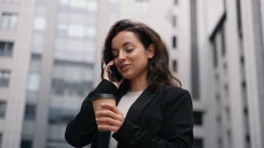 Caucasian female standing outdoors in the city center around offices and modern grey buildings. Young business woman talking on phone, smiling, looking around, having cup of coffee in her hand.
