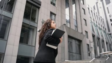 slow motion. Young Caucasian business woman stepping up to the office in the city center with high contemporary buildings. Girl running with a laptop, papers, and cup of coffee to go, looking around.