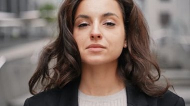 Close-up, slow motion. Young handsome woman looking into a camera, smiling, looking around, touching her dark long hair. Caucasian girl standing around grey modern building in the center of the city.