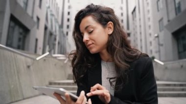 Beyaz iş kadını, şehir merkezindeki gri büyük binaların etrafındaki touchpad 'inde çalışıyor. Güzel kız gülümsüyor, daktilo ediyor, çalışıyor ve odaklanmış görünüyor. Uzak çalışma dijital uzmanı.