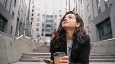 Working girl looking at her touchpad, and drinking beverage. Pretty young woman sitting on the stairs in downtown, drinking coffee, looking around, enjoying the view, and dreaming about the future.