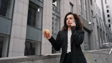 Caucasian woman staying on the stair, eating burger, talking on her phone, and working on the computer in the city center. A woman with brown long hair staying on stairs downtown working remotely.