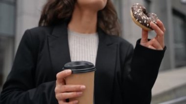 Young Caucasian girl eating donut and drinking coffee in the center of downtown. Woman with brown hair wearing black jacket having coffee and chocolate donut between skyscrapers. Outdoor, snacks time.