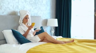 Caucasian beautiful female in white bathrobe and towel on her head is sitting on her bed in bedroom and texting on her phone. Pretty girl is drinking coffee and scrolling through her smartphone.