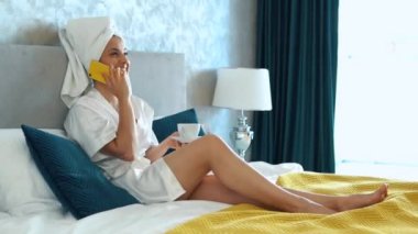 Cute Caucasian young woman in white bathrobe and towel on her head is sitting on her bed in bedroom and talking by phone. Pretty girl is laughing, smiling, enjoying conversation, drinking coffee.