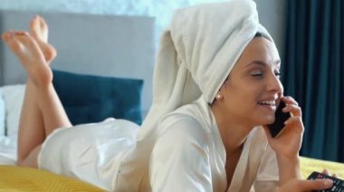 Cute Caucasian young woman in white bathrobe and towel on her head is lying on her bed in modern bedroom and talking by phone. Pretty girl talking, smiling, and enjoying conversation by phone.
