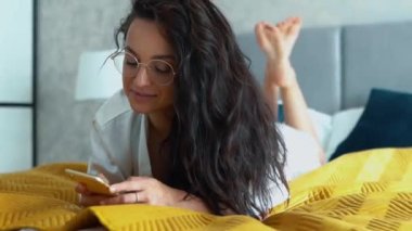 Caucasian pretty girl with long dark curly hair lying on bed, looking around and texting at her smartphone. Young woman in white t-shirt and glasses relaxing with pleasure on bed. Real-time closeup