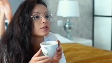 Caucasian sexy woman with long dark curly hair lying on bed, watching tv and drinking coffee from white cup. Girl in white t-shirt and glasses relaxing with pleasure on bed. Real-time closeup video.