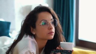 Caucasian pretty girl with long dark curly hair lying on bed, watching tv and drinking coffee from cup. Young woman in white t-shirt and glasses relaxing with pleasure on bed. Real-time closeup video.