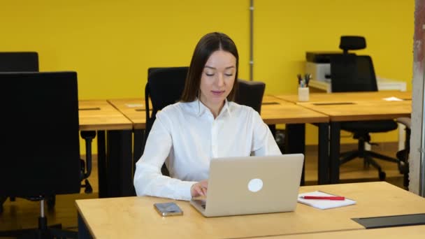 白いシャツを着た白人女性とグレーのズボンが現代の黄色のオフィスの彼女の作業台の上に座っている ビジネスマンの女性は彼女のコンピュータに取り組んでおり 非常に情熱的で真剣に見えます — ストック動画