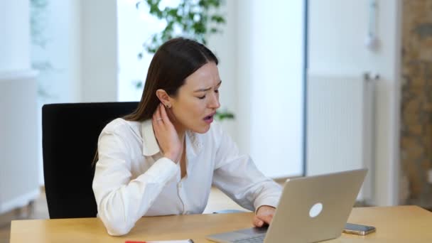 黒い髪の白人の若い女性は 彼女の首の痛みを満たす コンピュータ上で事務所で働いています 白いシャツを着たビジネスマンの女性がテーブルの上で働いていて 長い仕事の日の後にとても疲れています — ストック動画