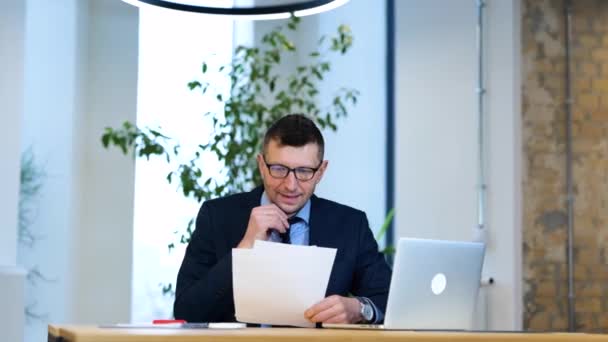 黒いスーツと青いシャツを着たビジネスマンがテーブルに座って 彼のコンピュータで作業し 書類を読んで 真剣に見ています オフィスで働いている白人男性は論文を読んで疲れている — ストック動画