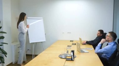 Beyaz kadın beyaz tahtaya diyagramlar çiziyor. İş kadını ofis alanında tahtaya yakın duruyor ve şirketin stratejisini erkek arkadaşlarına gösteriyor. Bunu tartışıyorlar..