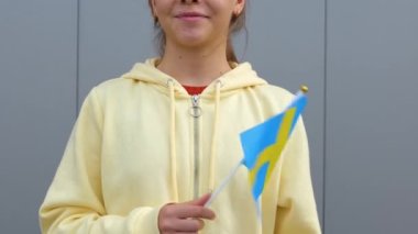 Beyaz, genç, güzel, sarı kapüşonlu kız küçük bir İsveç bayrağı sallıyor, kameraya gülümsüyor. Uzaklaş, yakınlaş, gri arkaplan, küçük kadın İsveç bayrağını elinde tutuyor..