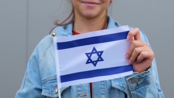 赤のTシャツとデニムのジャケットを着た白人の美しい十代の女の子は イスラエルの小さな旗を手に持っています 小さなイスラエル国旗を手に笑顔で手を振りながら外に立っています — ストック動画