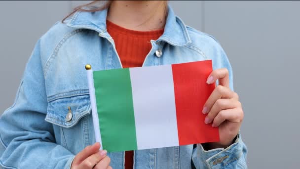 赤のTシャツとデニムのジャケットを着た白人の美しい十代の女の子は 彼女の手にイタリアの小さな旗を保持します 若い女性は屋外に立って 笑顔で手に小さなイタリアの旗を振っています — ストック動画