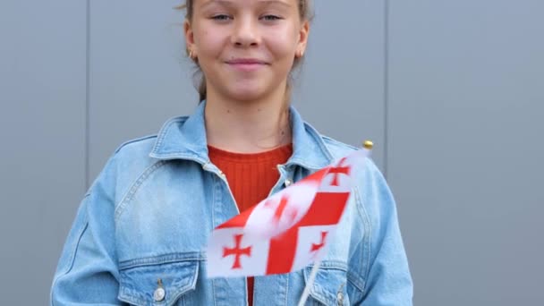 赤のTシャツとデニムのジャケットを着た白人の美しい10代の少女は ジョージアの小さな旗を手にしています 若い女性は屋外に立って笑顔と手に小さなグルジアの旗を振っている — ストック動画