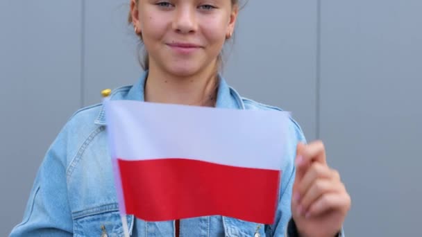 赤のTシャツとデニムのジャケットを着た白人ポーランドの10代の少女は ポーランドの小さな旗を手に持っています 若い女性は屋外に立って笑顔で手に小さなポーランド国旗を振っています — ストック動画