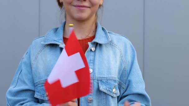赤のTシャツとデニムのジャケットを着た白人の10代の少女は スイスの小さな旗を手にしています 美しい若い女性は屋外に立っていて 笑顔で手に小さなスイスの旗を振っています — ストック動画