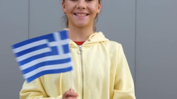 身穿黄色连帽衫的年轻白种人可爱的少女挥动着希腊的小国旗 对着镜头笑着 灰色背景 美丽的小女孩手里拿着希腊国旗 — 图库视频影像