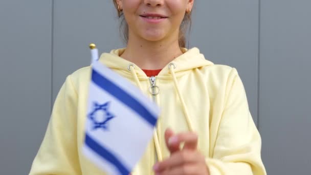 黄色のパーカーのかなり白人イスラエルの女性のティーンエイジャーは カメラで笑って 小さなイスラエルの旗を振っている ズームアウト 閉じる グレーの背景 小さな女の子は彼女の手にイスラエルのフラグを維持しています — ストック動画