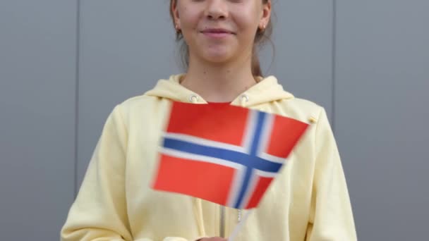 黄色のパーカーの白人の若い美しい10代の少女は カメラで笑みを浮かべて小さなノルウェーのフラグを振っている ズームアウト 閉じる 灰色の背景 小さな女性は彼女の手にノルウェーの旗を維持しています — ストック動画