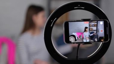Evde telefon kamerasıyla video kaydederken askılarda şık kıyafetler sergileyen neşeli genç bayan tripoddaki akıllı telefon ekranına odaklanıyor. Yavaş çekim videosu