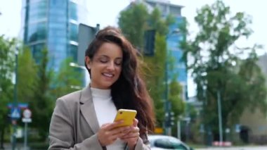 Sokakta dikilirken akıllı telefonu tutan mutlu kadın. İş kadını, mesaj yazıyor. Ofis binaları arka planda. Gerçek zamanlı.