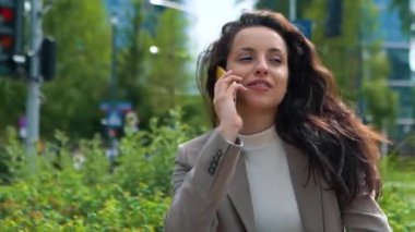Gülümseyen bayan, arka planda ofis binaları olan bankta otururken akıllı telefondan iş görüşmesi yapıyor. İş, teknoloji kavramı. Gerçek zamanlı.