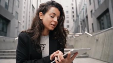 Dijital tablet kullanan ve arka planda ofis binaları olan dışarıda oturan genç bir bayan. İş kadını merdivenlerde oturuyor. İnternette çalışmak. Gerçek zamanlı.