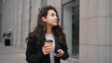 Mesajı okurken akıllı telefon kullanan genç bir bayan dışarıda iş merkezinin yanında duruyor. Elinde kahve tutan bir iş kadını. Yaşam tarzı, içki konsepti. Gerçek zamanlı.