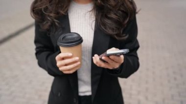 Güzel gülümseyen bayan dışarıda kahve içerken cep telefonunda bilgi buluyor. Ayakta duran ve telefonda konuşan bir kadın. Yaşam tarzı konsepti. Gerçek zamanlı.