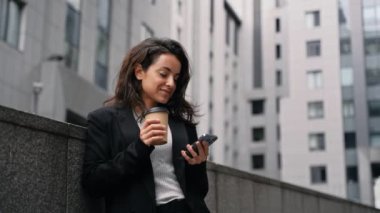 Mutlu güzel kadın duvarın yanında duruyor, kahve içiyor ve akıllı telefonda daktilo yazıyor. Bayan bakıyor ve ekrana vuruyor. Teknoloji, dijital konsept. Gerçek zamanlı.