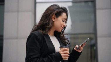 Güzel bir kadın dışarıda beklerken cep telefonu ekranından araklıyor. Kahve içen mutlu iş kadını. Teknoloji, dijital konsept. Gerçek zamanlı.