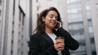 Gülümseyen bayan dışarıda duruyor, akıllı telefonla konuşurken tek kullanımlık kağıt bardakla kahveyi tutuyor. Arka planda iş merkezi var. Teknoloji, dijital konsept. Gerçek zamanlı.