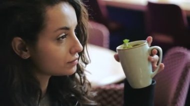 Lokantada oturmuş nane çayı içerken eğlenen mutlu, genç ve çekici bir kadın. Kahve dükkanında ferahlatıcı çay içen bir bayan. Antioksidanlı yeşil bitkiler. Yaşam tarzı konsepti. Gerçek zamanlı.