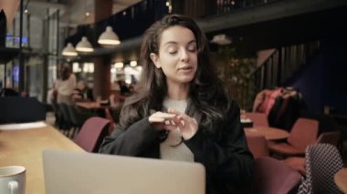 Kafede otururken kameraya bakıp konuşan mutlu genç bir kadın. Güzel bayan kafeteryada video görüşmesi yapıyor ve blog kaydediyor. Kafe 'de bir iş yerinde. Gerçek zamanlı.