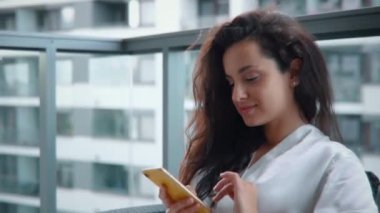 Evdeki balkonda elinde cep telefonuyla mesaj yazan mutlu bir bayan. İpek sabahlıklı kadın. Sabahın tadını çıkarma kavramı. Yaşam tarzı. Gerçek zamanlı.