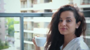 Beyaz sabahlık giymiş, balkonda elinde bir fincan kahveyle oturan mutlu genç bir kadın. Bayan dizüstü bilgisayar kullanırken çalışıyor. İletişim ağı, tele-çalışma konsepti. Gerçek zamanlı.