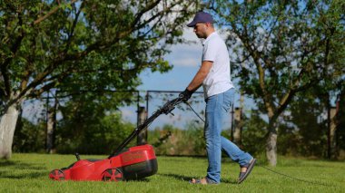 Yakışıklı erkek işçilerin özel bahçe çimleri biçerken paten makinesiyle yan görünümü. Adam bahçede yürüyor ve çim biçme makinesiyle yeşil çimleri biçiyor. Bahçıvanlık, bahçe bakımı. Yaz günü