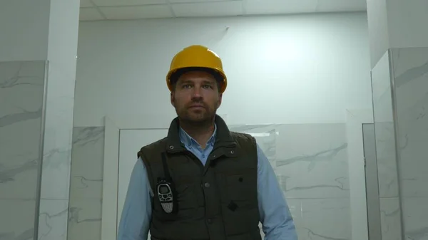 Close up portrait of young Caucasian male constructor in yellow helmet walks in building taking walkie-talkie on construction site. Handsome male architecture inspector in hard hat indoor. Slow motion
