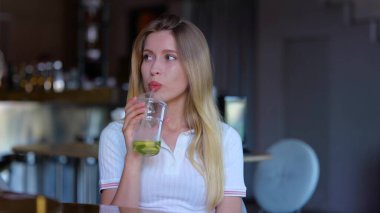 Close up of joyful young beautiful woman sitting in cafeteria and drinking ice tea. Pretty joyful Caucasian female with lemonade drink in restaurant indoors. Slow motion. Leisure. Drink concept