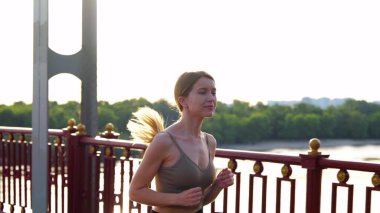 Portrait of beautiful positive female exercising outdoor on the bridge at sunrise in the morning. Healthy active lifestyle. Happy young woman running outside in city on summer morning. Workout concept