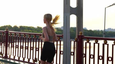 Back view of Caucasian young slim woman running on street in town on the bridge. Rear of fit female in sportswear runs outdoor. Healthy lifestyle. Slow motion. Sporty concept. Sport workout outside