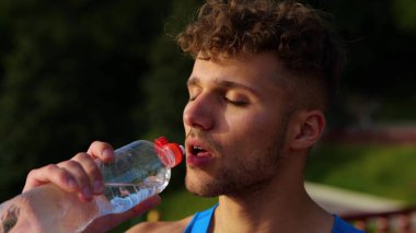 Handsome young sportsman with serious face drinking water from bottle while standing outdoors after workout. Caucasian athletic man with curly hair drinks water on sunny day. Sport activity. Close up