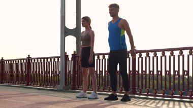 Camera approaching young couple of man and woman in sportswear working out outside on the bridge. Caucasian sporty male and female doing exercise outdoor. Athletes doing squats. Sport activity.