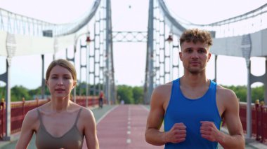 Close up of Caucasian joyful young man and woman friends running working out outdoors. Male and female jogging on beautiful bridge together. Sport activity. Healthy life. Couple workout concept