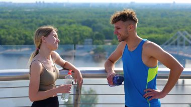 Close up portrait of joyful Caucasian young sportswoman speaking with handsome sportsman outside in city with bottle of water in hands. Male and female athletes laughing and chatting after working out